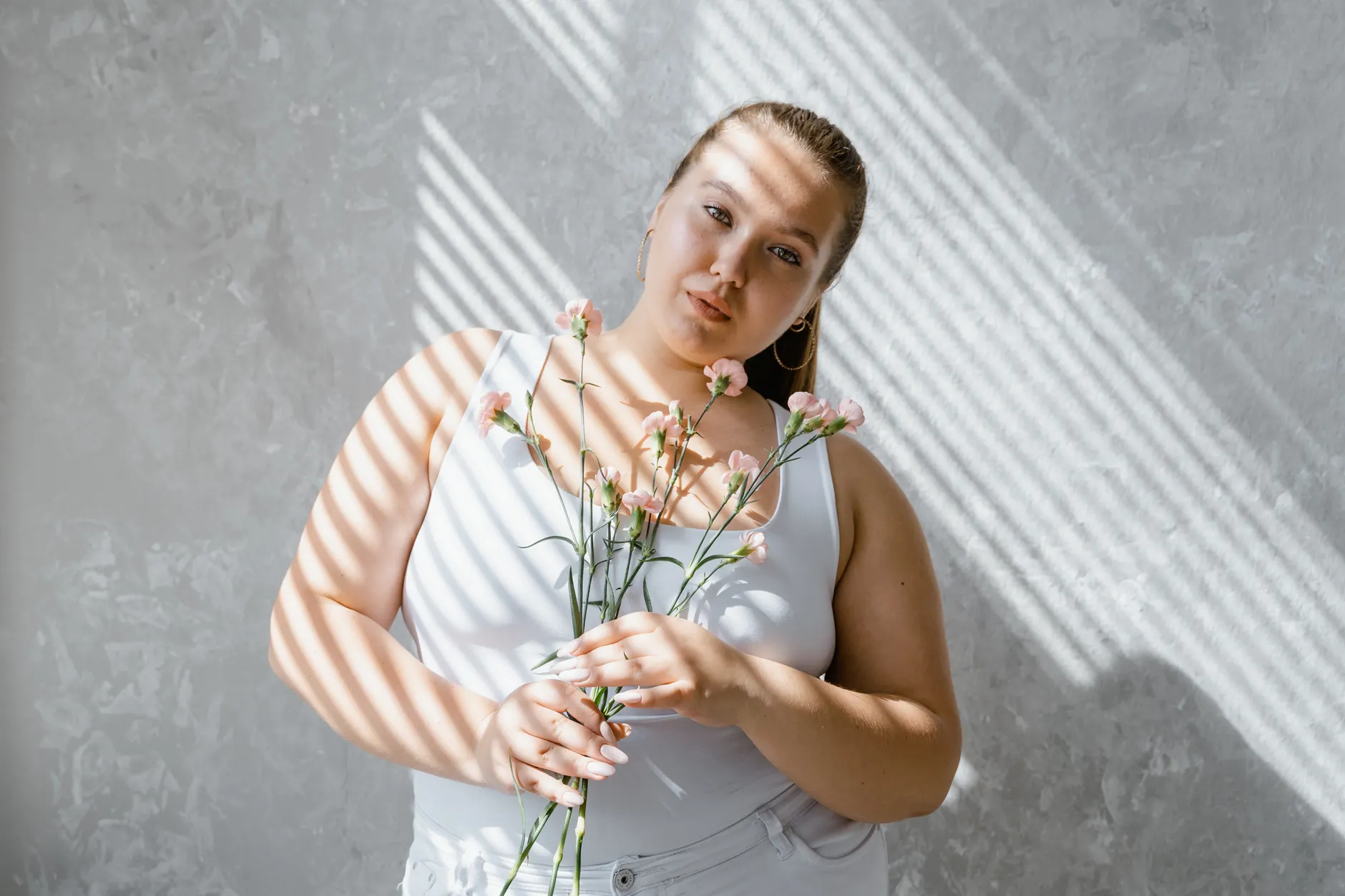 Plus-size woman in tank top holding flowers with sunlight filtering through blinds.
